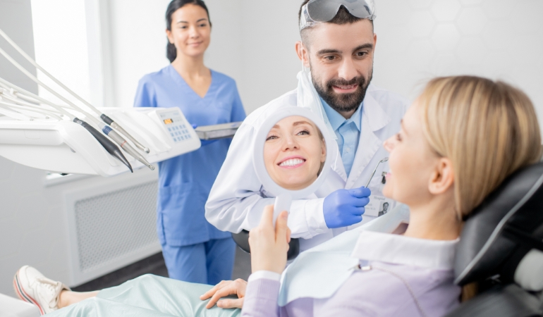 young-smiling-woman-looking-mirror-after-procedure-professional-whitening-while-sitting-armchair-dentist-office